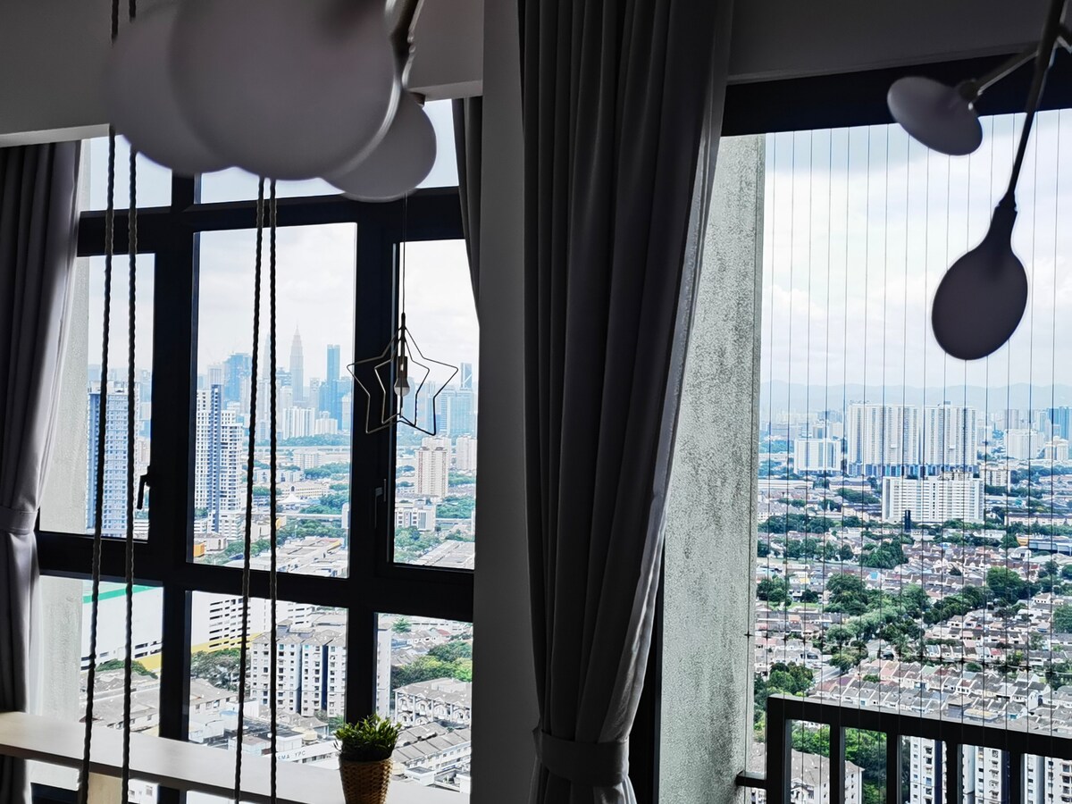A large window spans the wall, offering a panoramic view of the city skyline. Tall buildings and greenery can be seen in the distance. Soft gray curtains frame the window, while modern pendant lights hang above a dining area, adding to the overall ambiance.