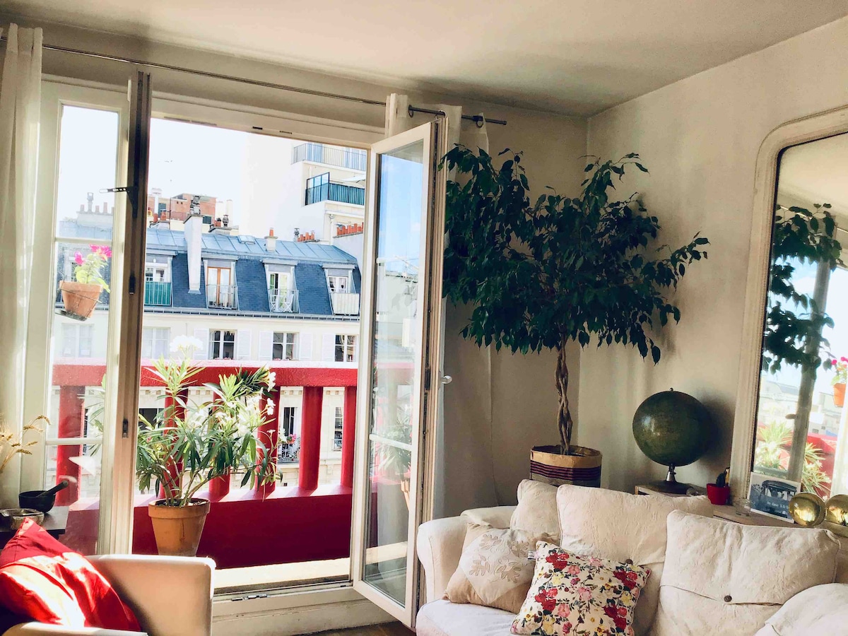 A bright living area is visible, featuring large glass doors that open to a balcony with a view of Parisian rooftops. A potted plant and decorative globe add character to the space. Light filters through the windows, enhancing the tranquil atmosphere.