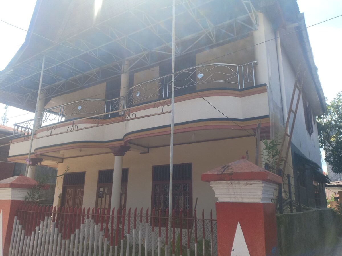 The exterior of a two-story building is presented, featuring a covered balcony with decorative railings. A wall is seen in front, painted in red and white, enclosing a garden area with greenery. The structure's architecture reflects local design elements.