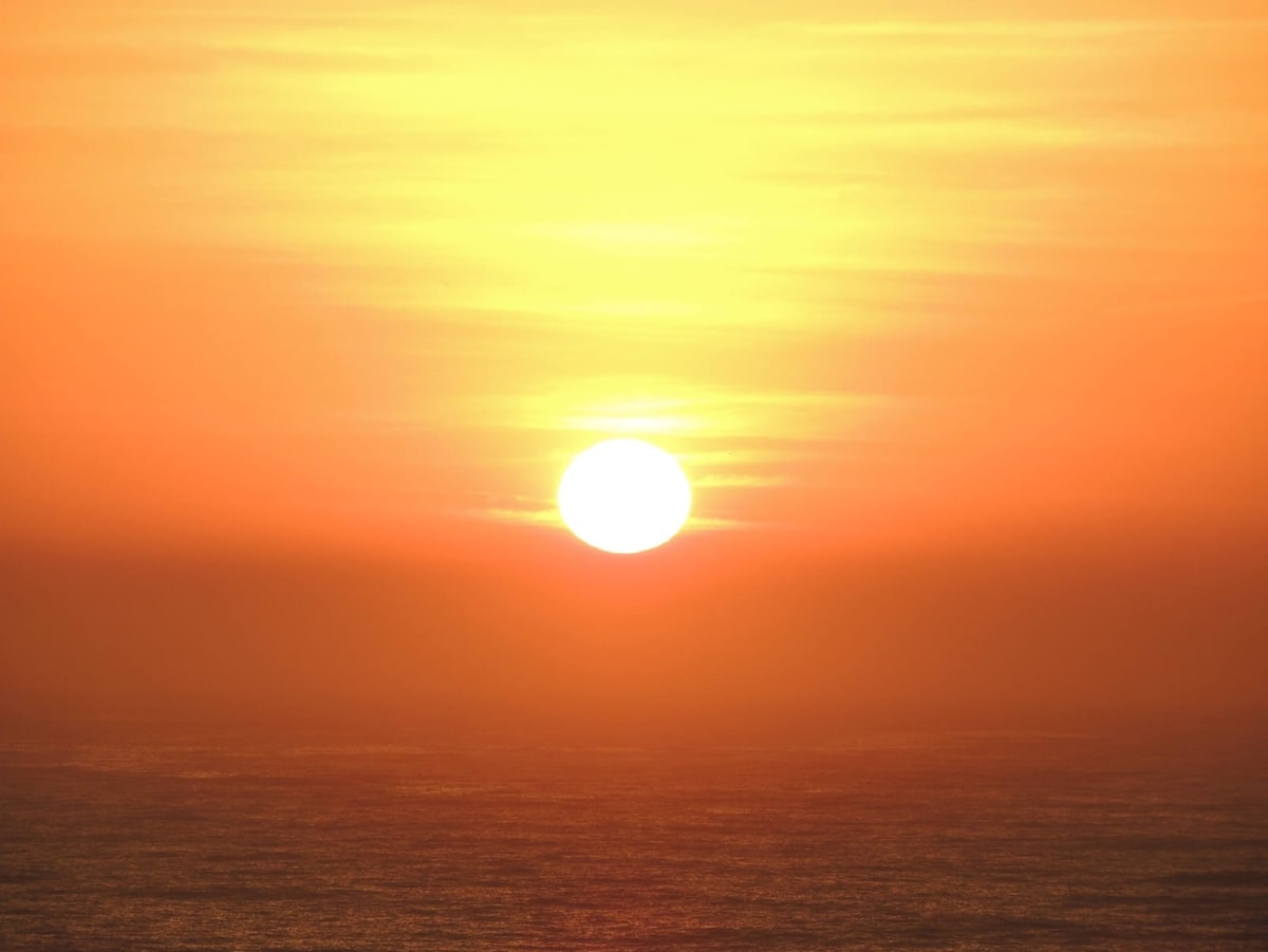 A vivid sunset is captured over the horizon, with the sun casting a warm orange glow across the ocean's surface. Soft waves are visible below, reflecting the evening light.