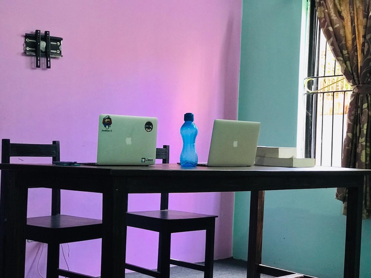 A wooden table is set in a workspace with two laptops positioned on it. The background features a pastel purple wall paired with a teal section. Natural light enters through a window adorned with patterned curtains, enhancing the bright and functional atmosphere.