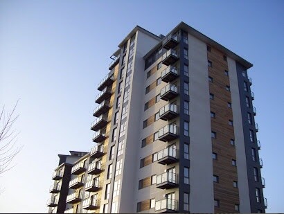 A modern apartment building rises against a clear blue sky, showcasing a combination of glass balustrades and wooden accents. Multiple balconies are visible on the facade, providing outdoor space for upper-floor residents.