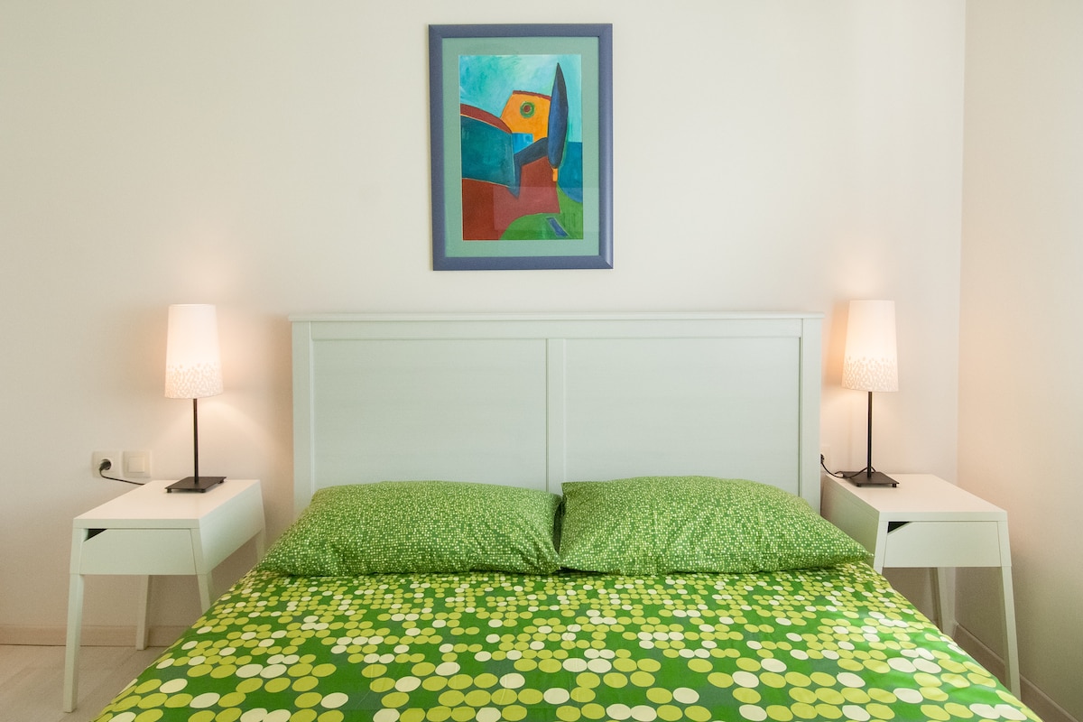 A cozy sleeping area features a large headboard framing a double bed with vibrant green bedding adorned with circular patterns. Flanking the bed, two bedside tables hold softly glowing lamps. A colorful artwork hangs above the bed, enhancing the serene atmosphere.