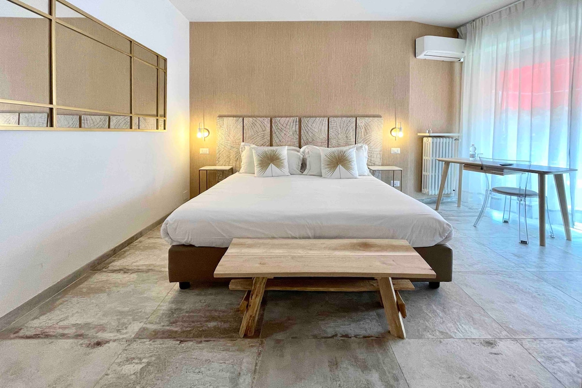 A spacious sleeping area is highlighted by a large bed with a neutral-toned headboard. Warm lighting fixtures flank the bed, illuminating the room. A wooden bench is placed at the foot of the bed, and a glass dining table is visible near a large window with sheer curtains.