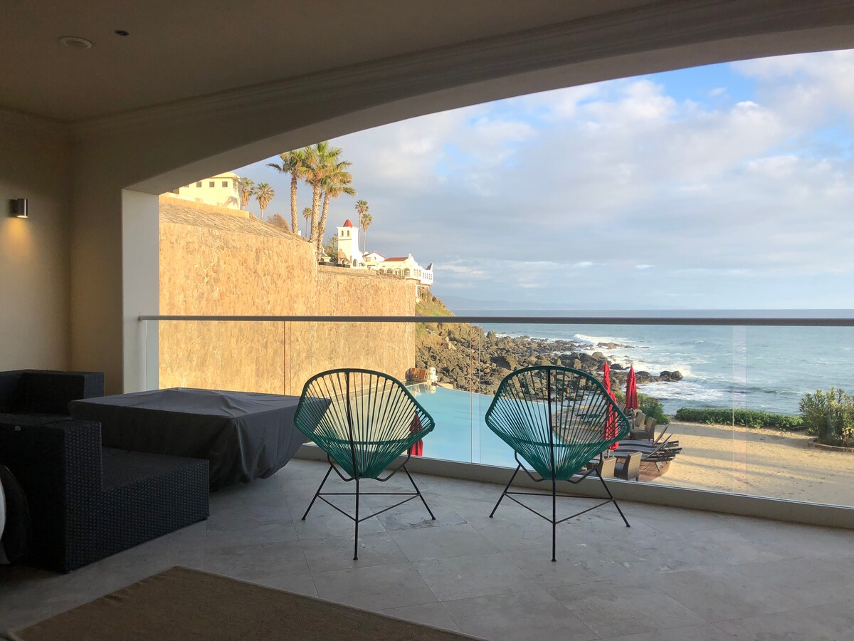 The image captures a serene balcony area with two Acapulco chairs facing an ocean view. A stone wall and palm trees create a natural backdrop, while waves gently meet the shore. A covered seating arrangement is visible to the left, adding to the inviting atmosphere.