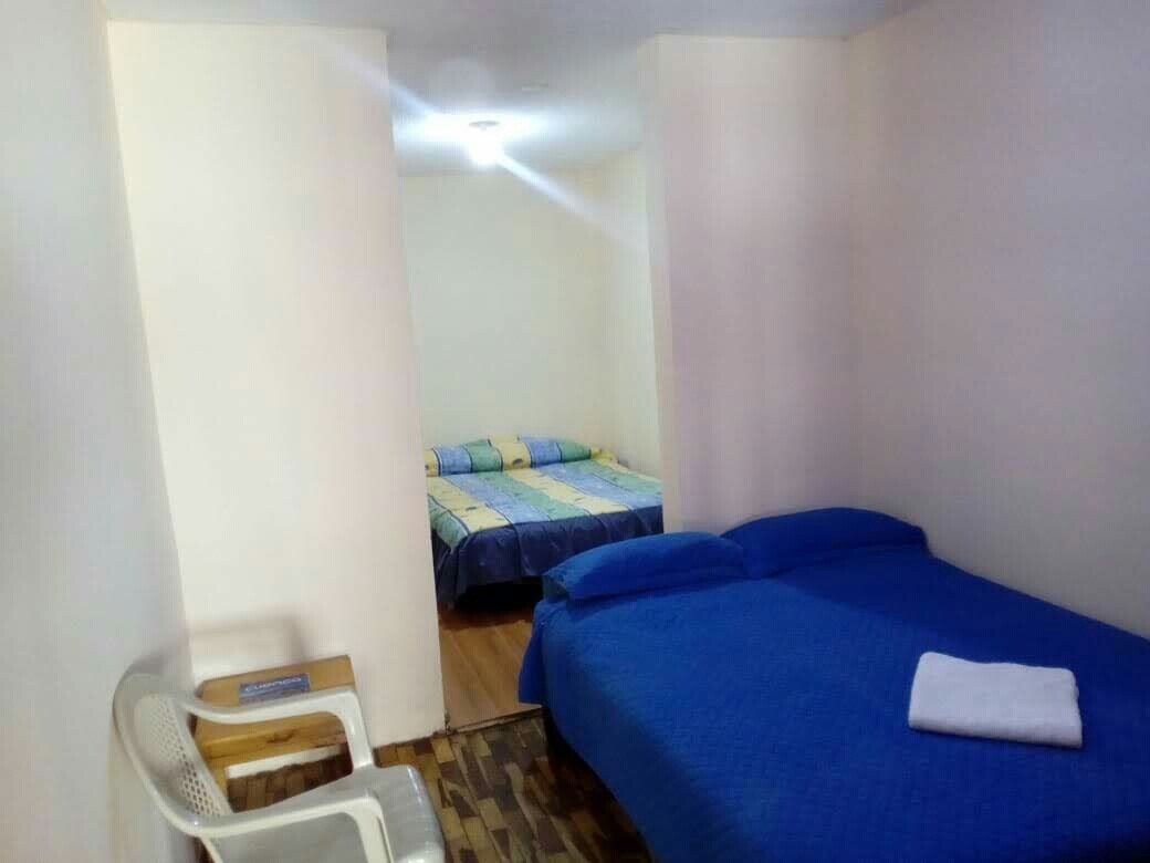 A bedroom is shown with two beds, one larger and one smaller, each made with blue bedding. A white towel is neatly placed on the larger bed. A plastic chair is positioned next to the larger bed, and light is provided by a ceiling fixture.