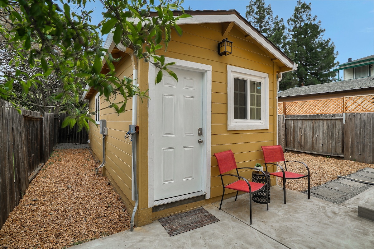 The exterior of the studio is highlighted by its cheerful yellow siding. A white door provides access, flanked by two red chairs and a small round table. The surrounding area features a gravel pathway and wooden fencing, enhancing privacy.