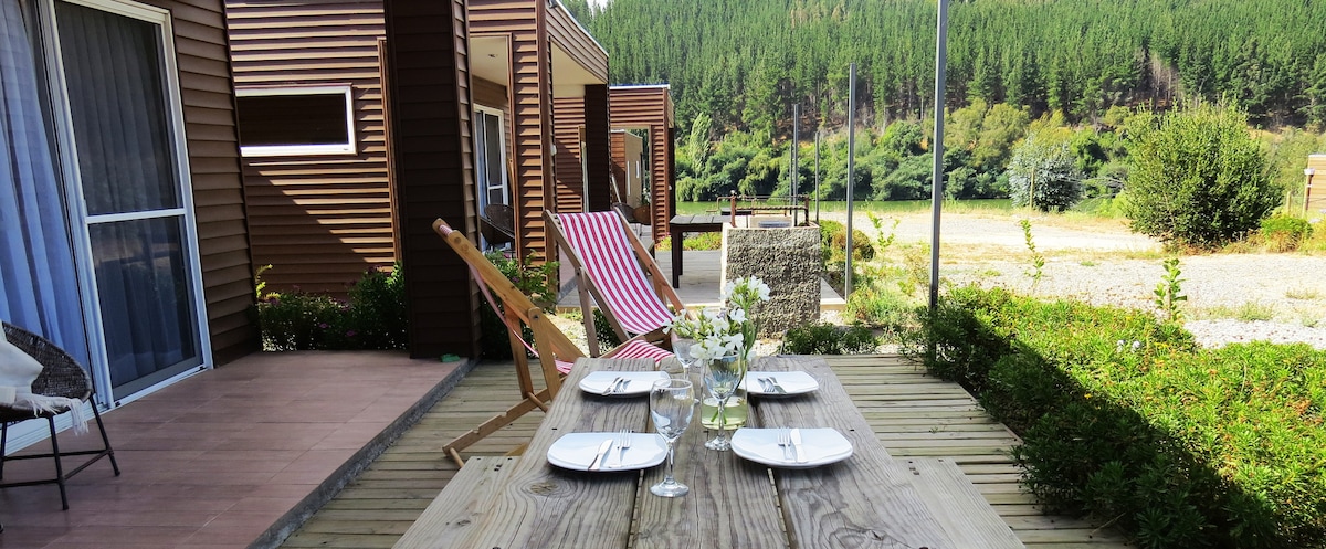 An outdoor dining area is set with a wooden table, featuring plates and glasses. A folding chair and chaise lounge with striped fabric are positioned nearby. The backdrop includes lush green hills and a clear view of the surrounding nature.