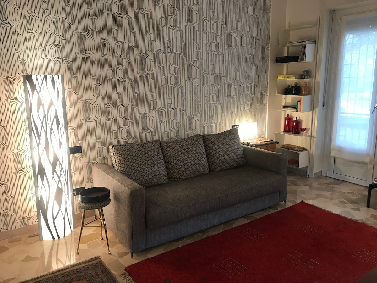 A comfortable gray sofa is positioned against a textured wall, accompanied by a small side table. A stylish floor lamp casts a soft light, enhancing the cozy ambiance. A red area rug adds warmth to the space, which is accented by a small bookshelf holding three visible objects.