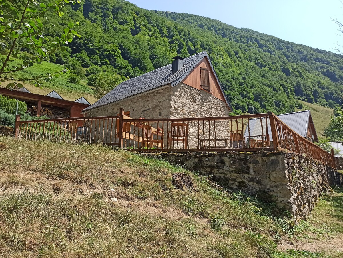 A charming chalet is set amidst lush greenery, featuring a stone exterior and a sloping roof. A wooden terrace furnished with several chairs offers an inviting outdoor space, while the backdrop displays the surrounding verdant hills.