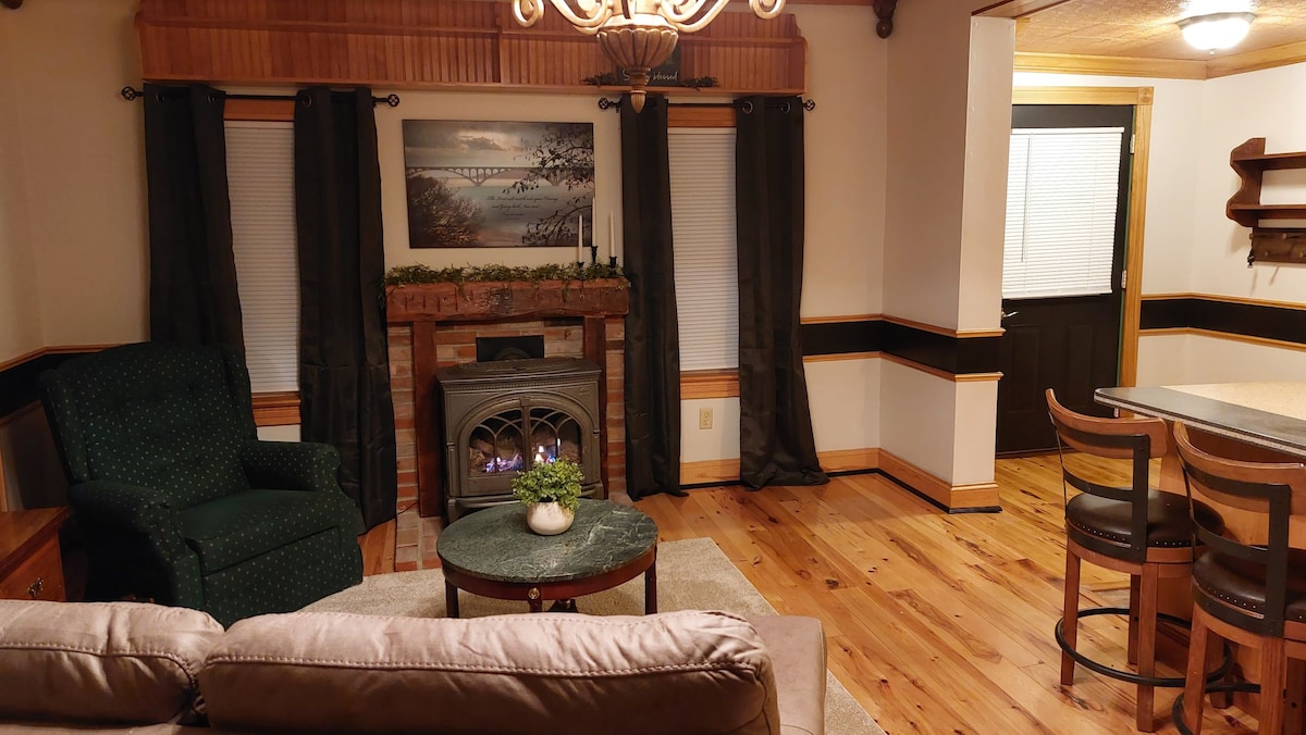 The cozy living area features a comfortable green armchair and a beige sofa. A circular table sits in the center, with a small potted plant displayed on top. A fireplace is framed by a stone mantel, and dark curtains cover the windows, providing a relaxing ambiance.