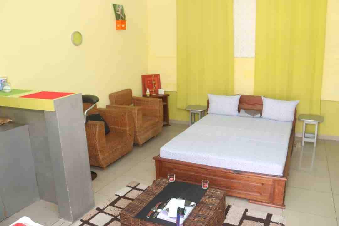 A cozy room features a spacious bed with light-colored bedding, complemented by a woven coffee table. A pair of rattan chairs and a small table are positioned nearby. Bright yellow curtains frame the window, enhancing the room's cheerful ambiance.