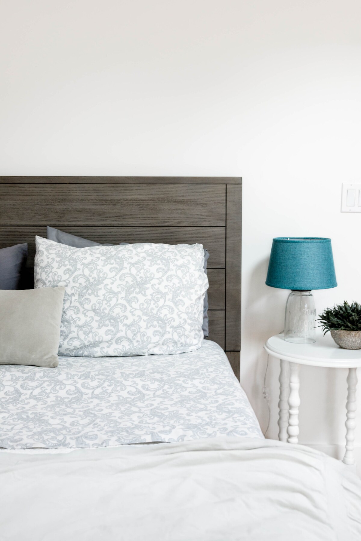 The bedroom features a neatly made bed with light grey and white patterned bedding. A decorative pillow and a soft cushion rest on the bed. Beside it, a small round table holds a teal lamp and a potted plant, adding a touch of comfort.