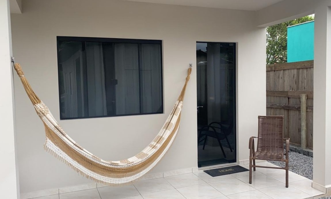 A cozy outdoor area features a light-colored hammock suspended against a smooth wall. A dark paneled window allows natural light to enter, and a brown wicker chair is positioned nearby on the tiled flooring. A simple welcome mat is placed at the entrance.