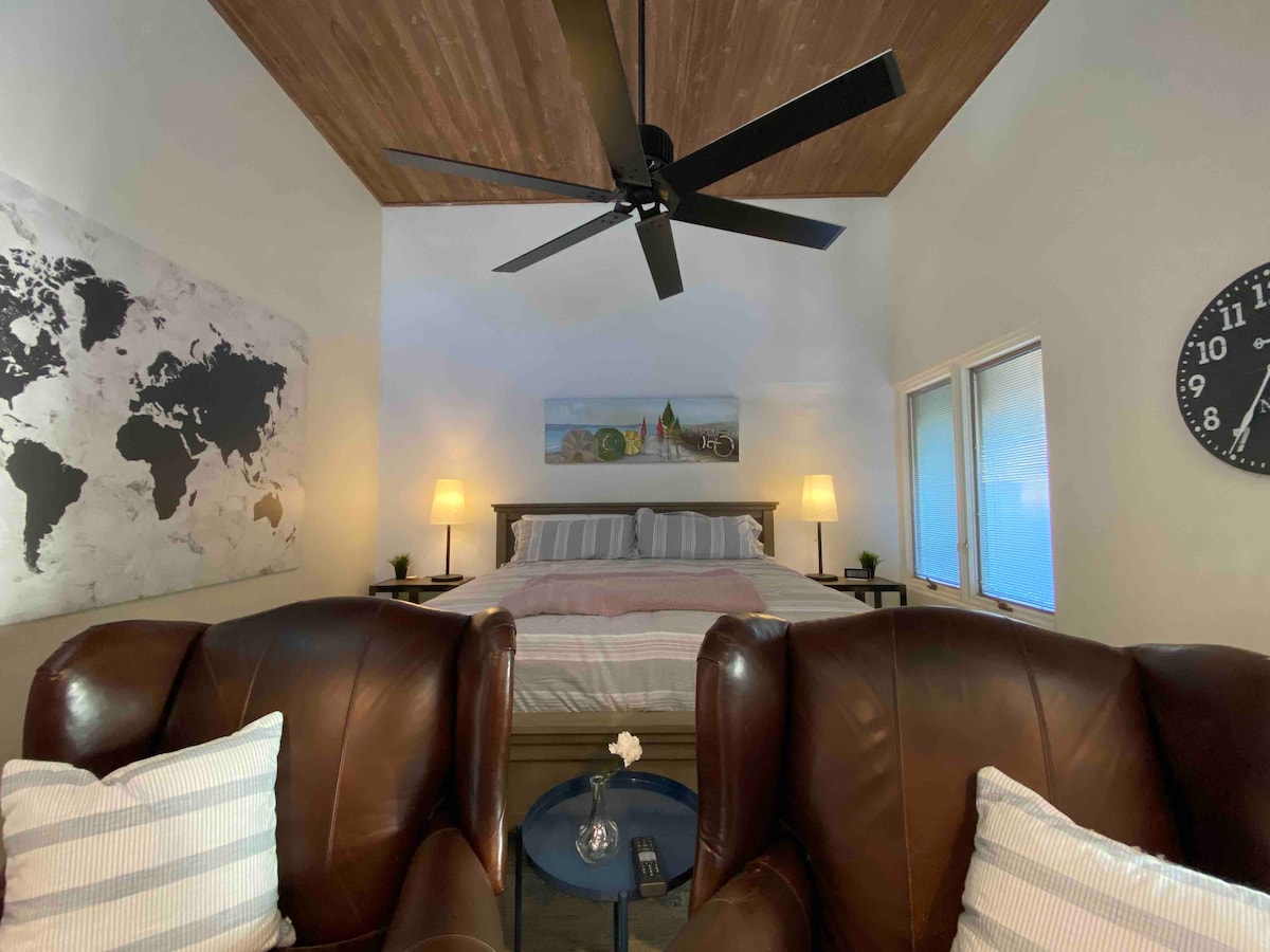 A cozy sleeping area features a spacious bed with soft linens and two bedside lamps. Brown leather chairs are positioned in the foreground, enhancing comfort. A world map adorns the wall, and large windows allow natural light to fill the space, showcasing a wooden ceiling.