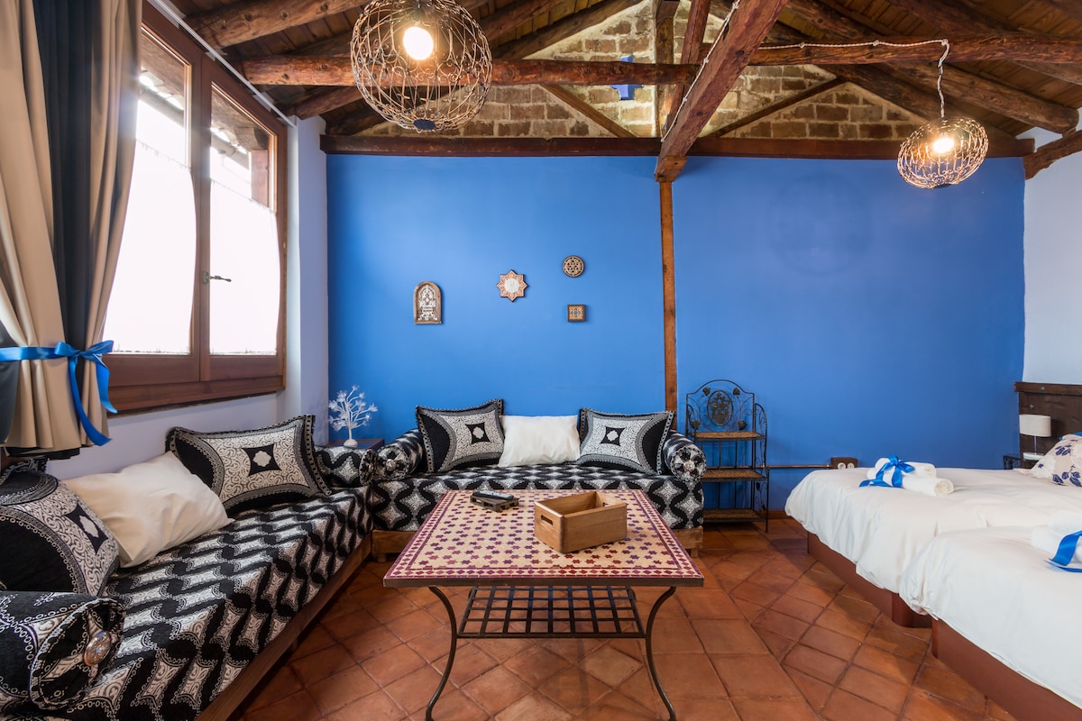 A vibrant lounge area features two sofas adorned with black and white patterns, accompanied by a central table with a colorful design. Exposed wooden beams accentuate the ceiling, and large windows allow natural light to fill the space.