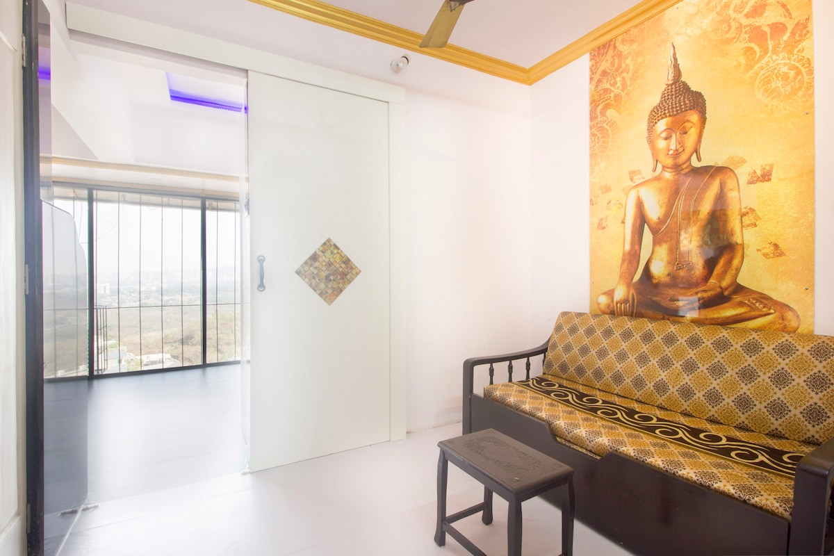 A serene corner of the apartment features a cozy seating area with a patterned couch and a small black table. A mural of a Buddha figure adds a calming element to the space. Large glass doors provide a view of the outside landscape.