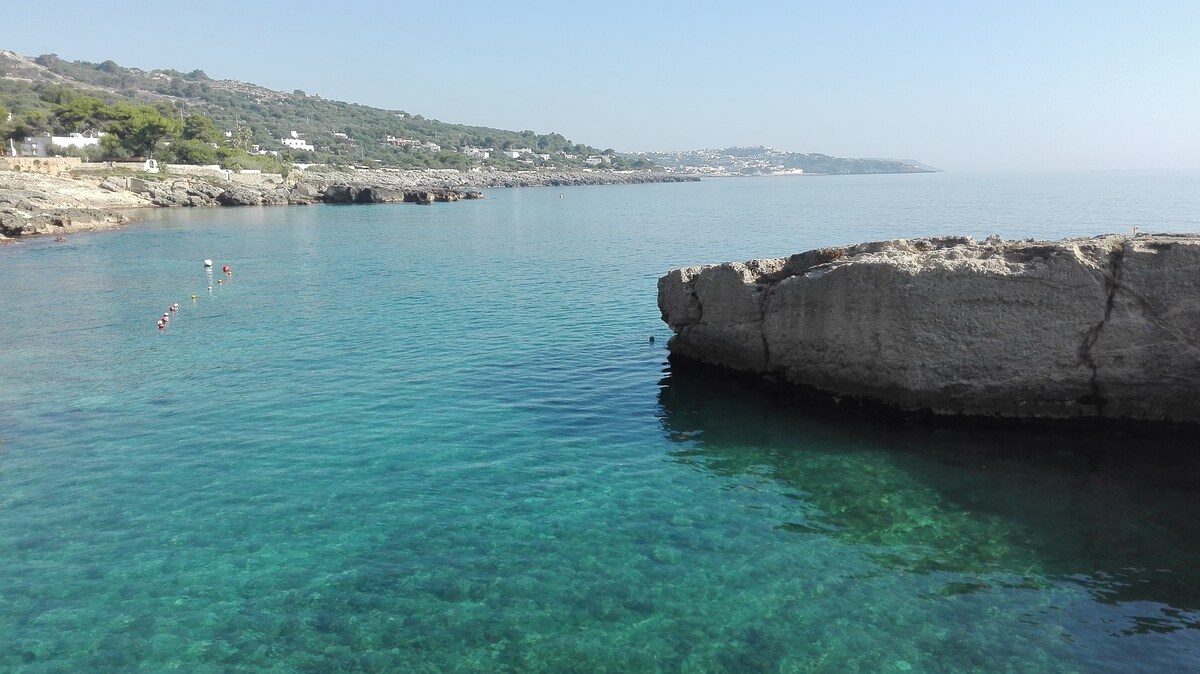A calm, clear sea reflects the blue sky in a tranquil coastal setting. Smooth rock formations are visible along the shoreline, with gently rolling hills in the background. Nearby, a few individuals can be seen wading in the shallow waters, enjoying the serene atmosphere.