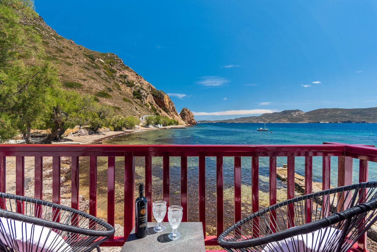 A balcony overlooks the serene waters, featuring two cozy chairs positioned around a small table. Glasses sit elegantly on the table, reflecting the sunlight. The scenic backdrop includes distant hills and a clear blue sky, creating a peaceful coastal view.