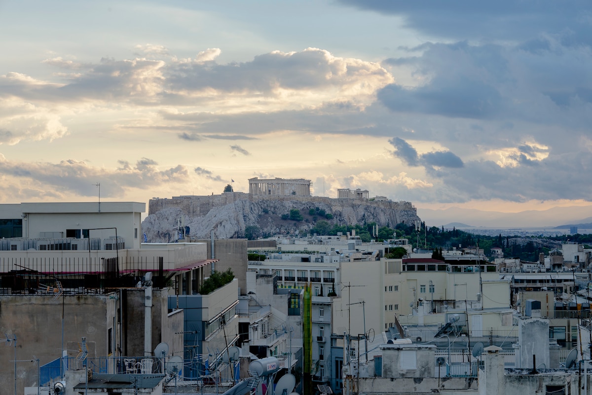 A panoramic view of the Acropolis is captured at sunset, highlighting its historic architecture against a backdrop of gently rolling clouds. The rooftops of surrounding buildings are visible in the foreground, creating a contrast with the ancient site perched atop the hill.