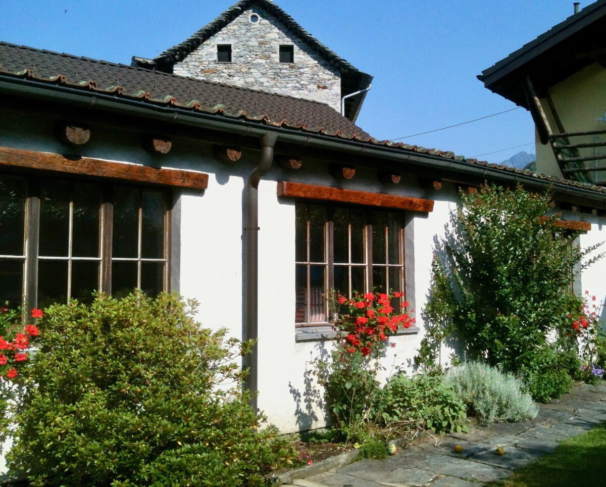 The exterior of a rustic house is shown, featuring a combination of stone and wooden elements. Colorful flowers and greenery surround the windows, enhancing the charming appearance. A clear blue sky complements the inviting atmosphere of the peaceful garden.