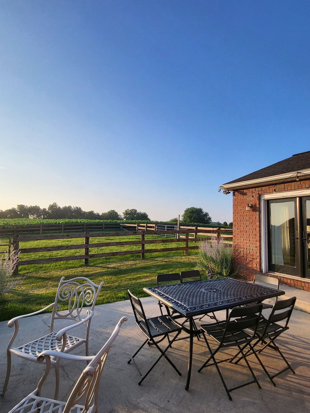 An outdoor patio area features a black table surrounded by folding chairs. A well-maintained lawn extends beyond a wooden fence, leading to open farmland under a clear blue sky. Light casts gentle shadows across the concrete surface, enhancing the tranquil setting.
