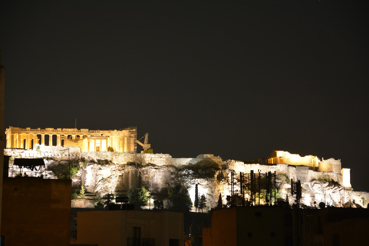 The illuminated Acropolis can be seen against the night sky, showcasing its ancient structures bathed in warm yellow light. The surrounding landscape is dark, providing a striking contrast to the bright, historic site atop the hill.
