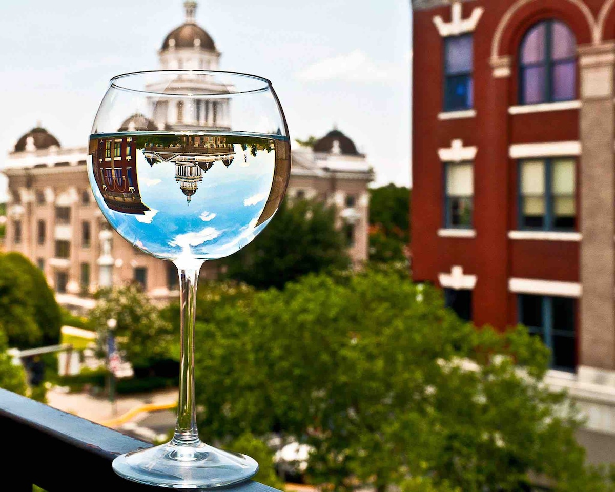 A large wine glass filled with clear water is placed on a ledge, reflecting the exterior of a historic building in the background. Lush greenery surrounding the glass enhances the serene setting, creating a harmonious view of the architecture.