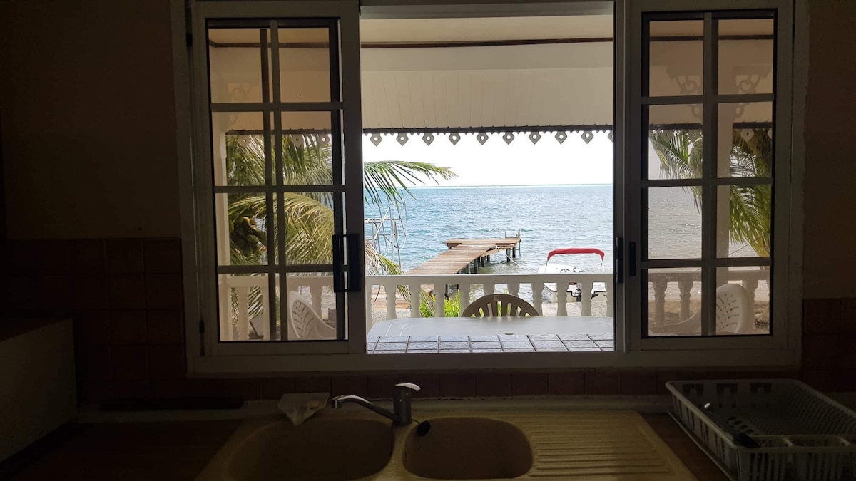 A view from the kitchen features large open windows overlooking the serene sea. A wooden pontoon is visible just beyond the shore, surrounded by palm trees. The scene invites natural light into the space, creating a seamless connection with the outdoor environment.