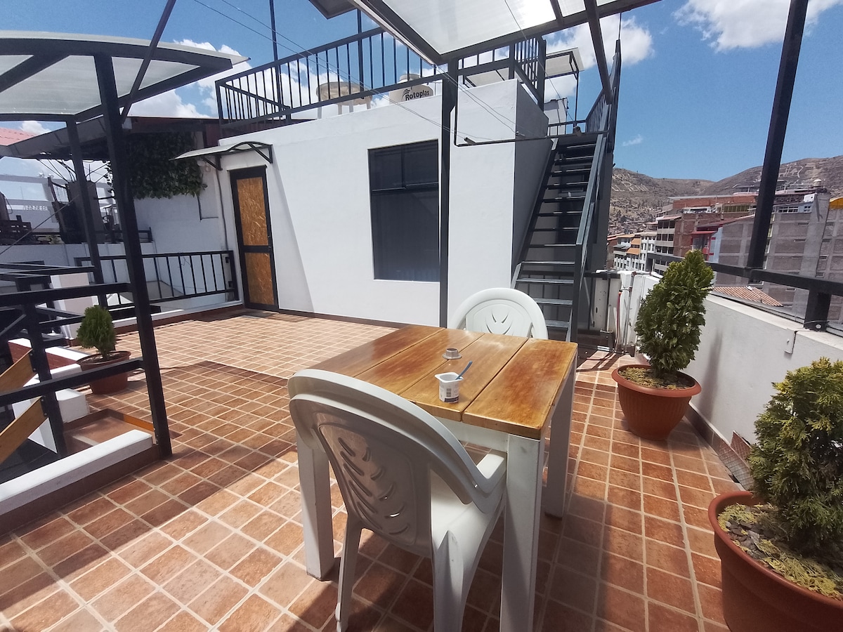 A rooftop terrace is showcased, featuring a wooden table with four plastic chairs. Potted plants are positioned around the area. The view extends to the surrounding buildings and mountains under a clear blue sky. A staircase leads to another level, adding accessibility.