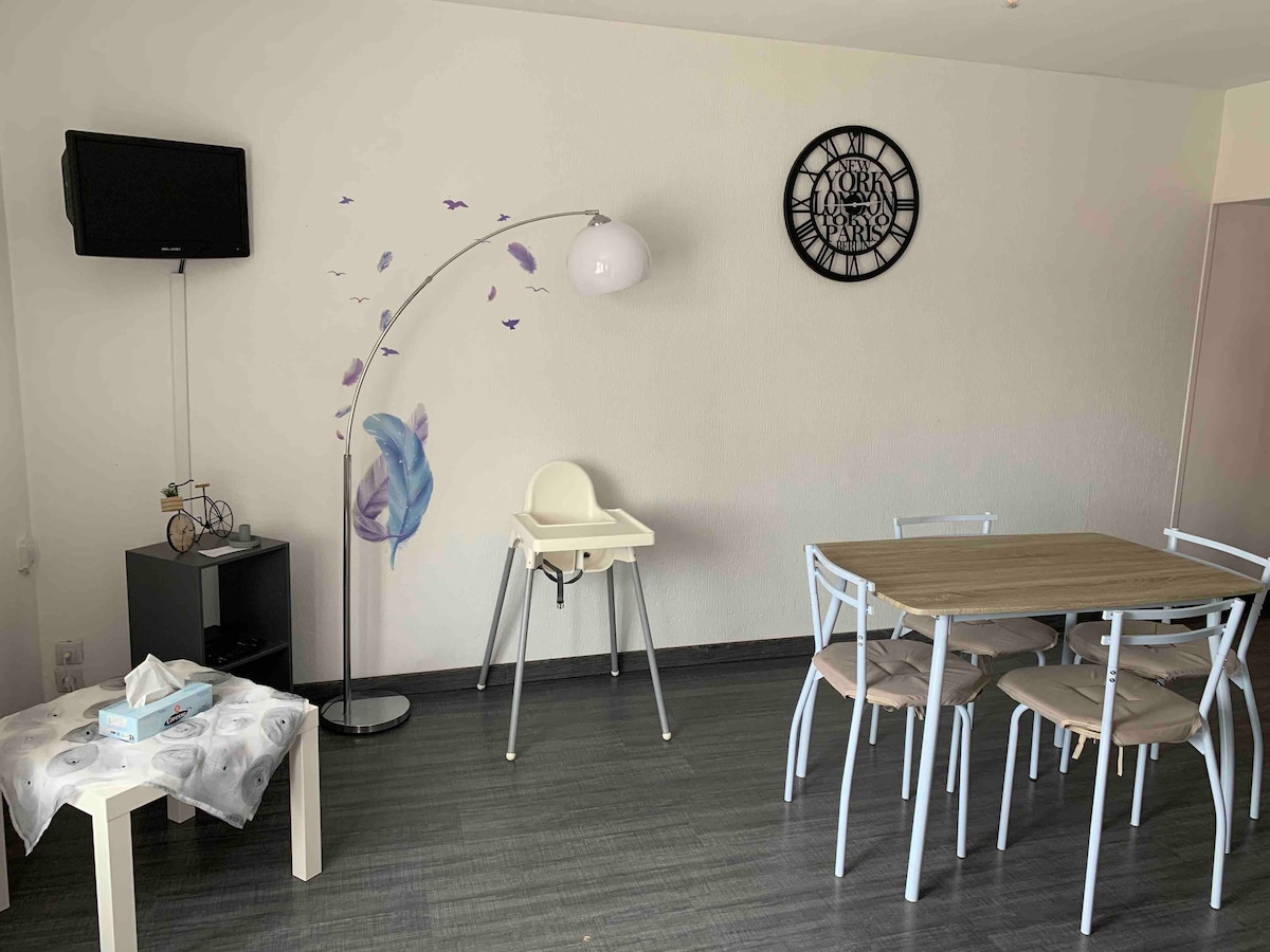 A light-filled living area features a wooden dining table surrounded by four light-colored chairs. A high chair is positioned nearby. A standing lamp provides additional lighting, and a television is mounted on the wall while a decorative clock adds a stylish touch.