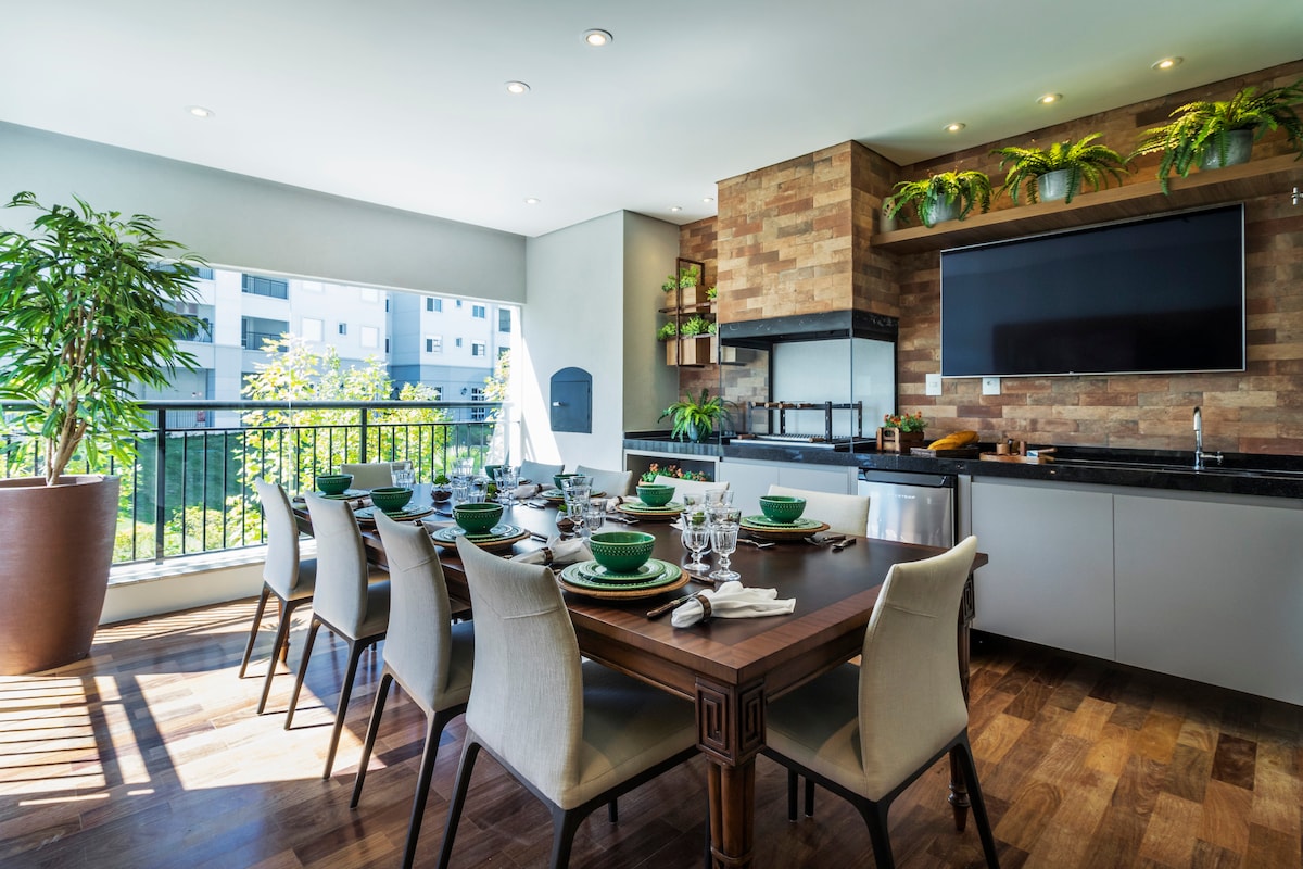 A spacious dining area features a large wooden table surrounded by light-colored chairs. Large windows allow natural light to fill the room, with greenery enhancing the atmosphere. A modern kitchen setup is visible, along with a mounted television and elegant dining ware on the table.