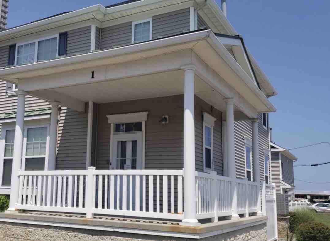 The exterior features a welcoming porch with white railings and pillars, providing an inviting entrance. Natural light enhances the light-colored siding of the house, while nearby views of the neighborhood and sky add to the overall ambiance.