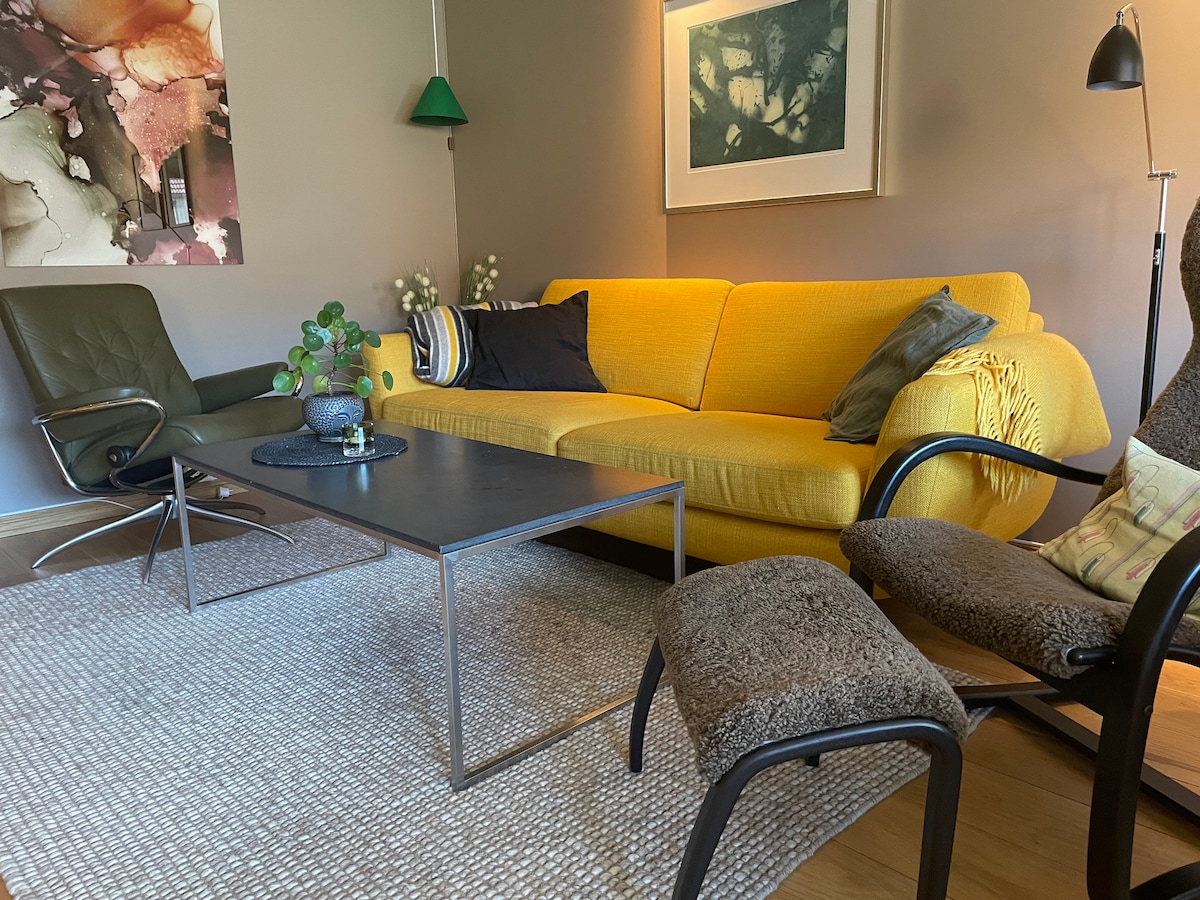 A bright yellow sofa is positioned in a cozy living area, accompanied by a dark green chair and two patterned armchairs. A square table and a small plant center the space, which is adorned with a warm area rug and wall art that adds character.