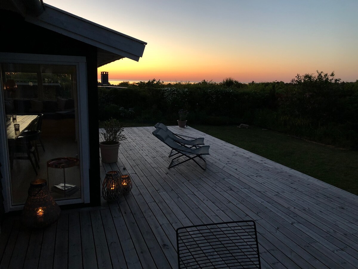 A wooden terrace is visible, featuring two lounge chairs positioned for sunset views. Soft lighting from lanterns creates a cozy ambiance. In the background, a serene sea can be seen under a colorful sky, while lush greenery surrounds the area.