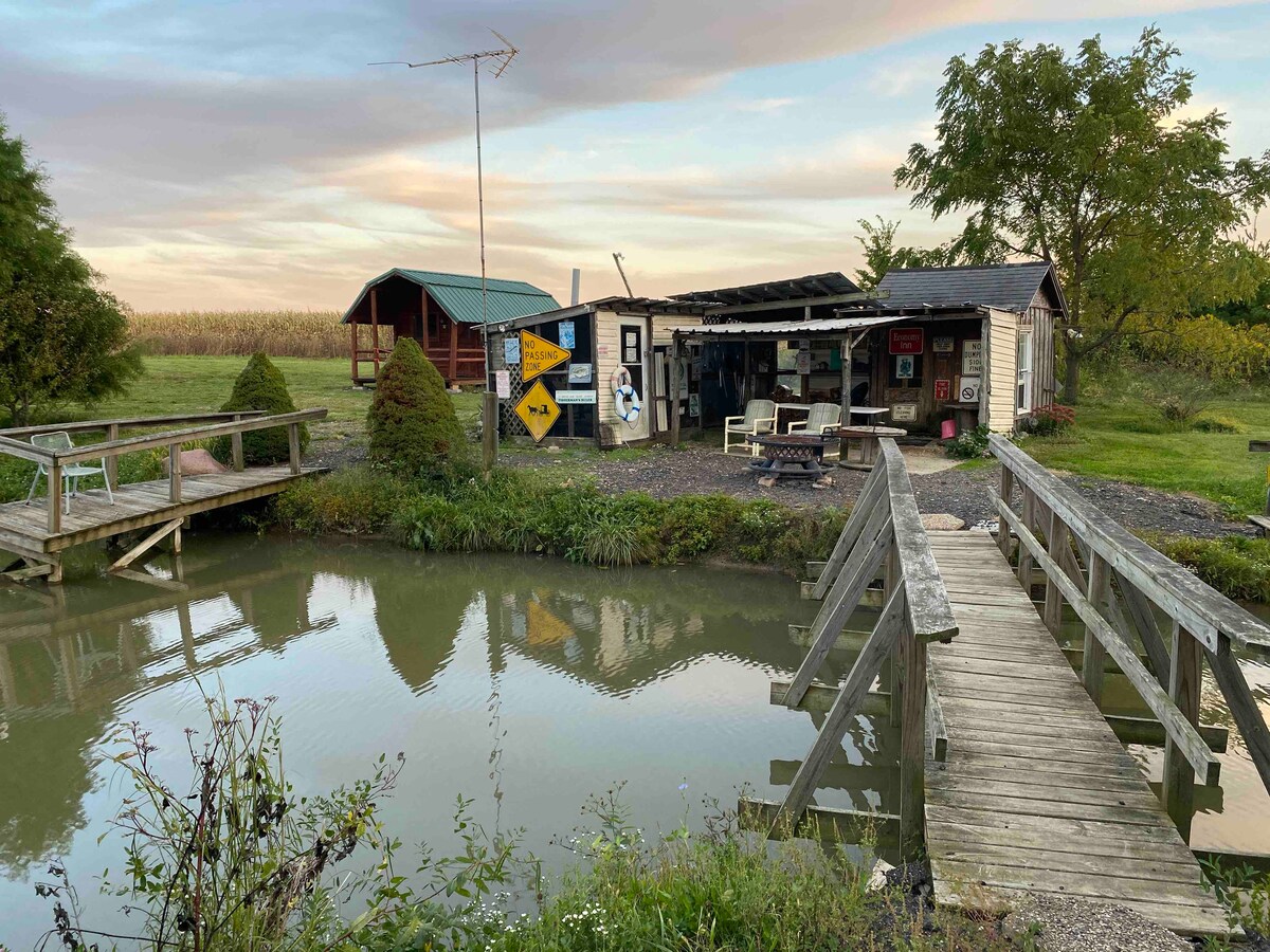 A rustic cabin and additional structure are visible beside a tranquil pond. A wooden bridge leads to the cabin, surrounded by greenery and a patch of grass. Signs and various items are arranged outside the shed, set against a backdrop of serene skies.