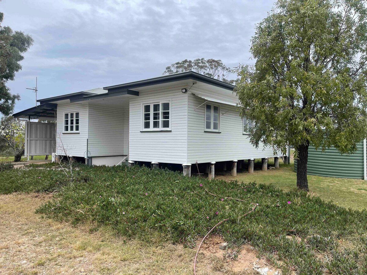 The exterior of the farm-house cottage is showcased, featuring a white clapboard design. The building is elevated on stilts, surrounded by a grassy area with shrubs. Large windows are visible, allowing natural light into the interior. A tree provides shade nearby.