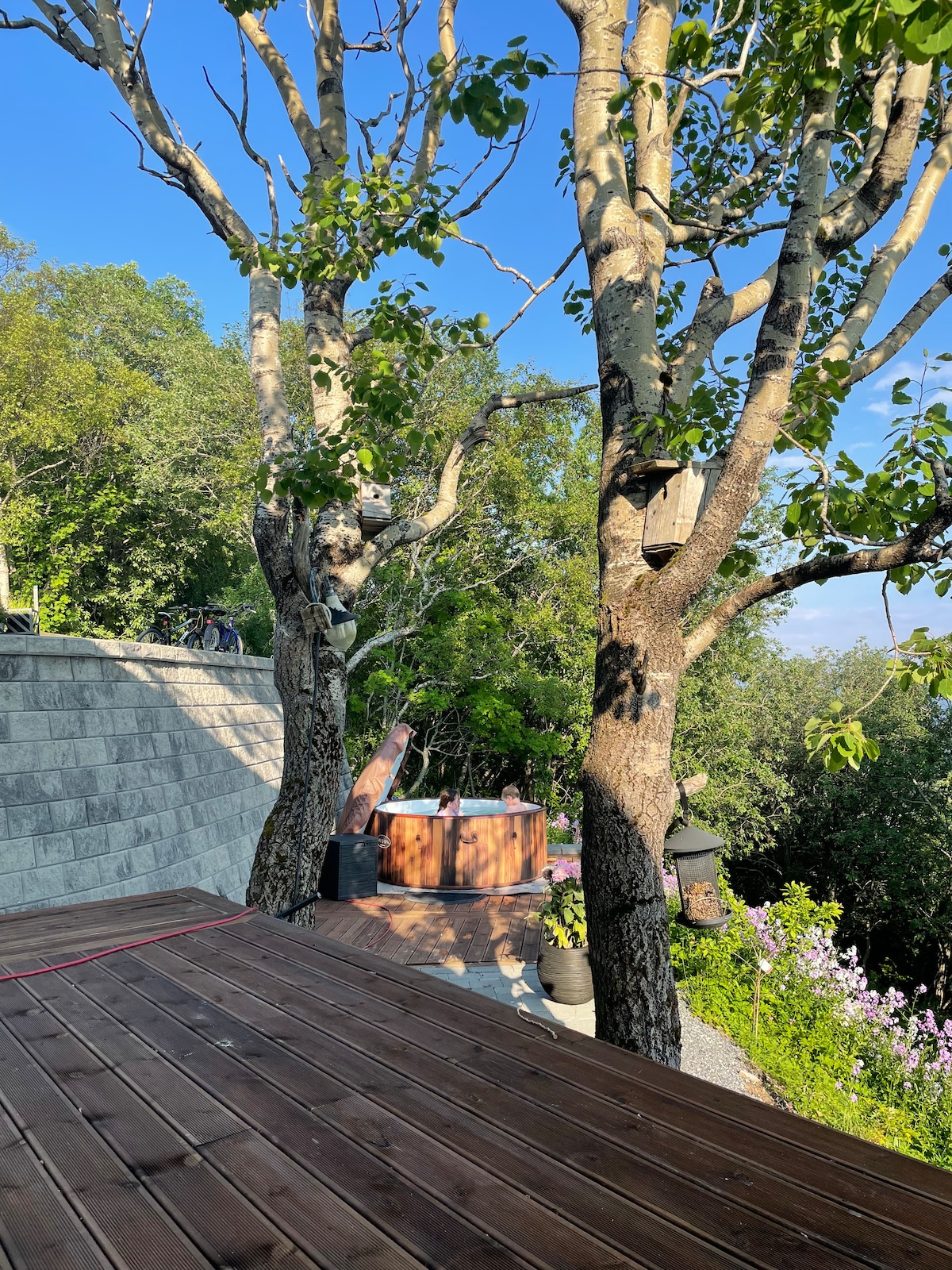 A wooden deck offers a view of lush green trees and a hot tub nestled among natural surroundings. The area features stone steps leading up to the home, with colorful flowers adding a touch of vibrancy to the scene.