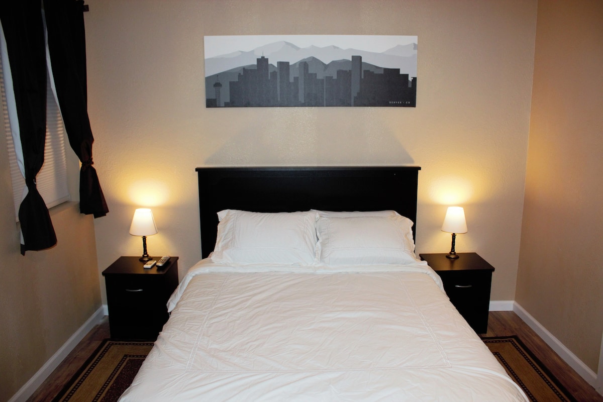 A cozy bedroom is shown, featuring a spacious bed with crisp white linens. Nightstands with lamps flank either side of the bed, casting a soft glow. A minimalist black and white cityscape artwork is displayed above the headboard, while dark curtains frame the window.