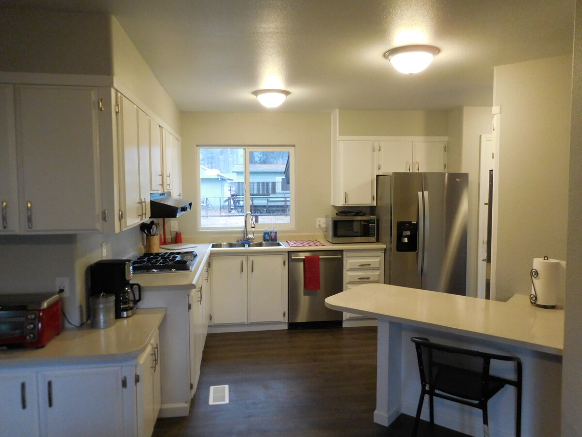 A modern kitchen features white cabinetry and stainless steel appliances, including a refrigerator, oven, and dishwasher. A window provides natural light and a view of the outdoor space. A small dining area with a chair is positioned near a breakfast bar.