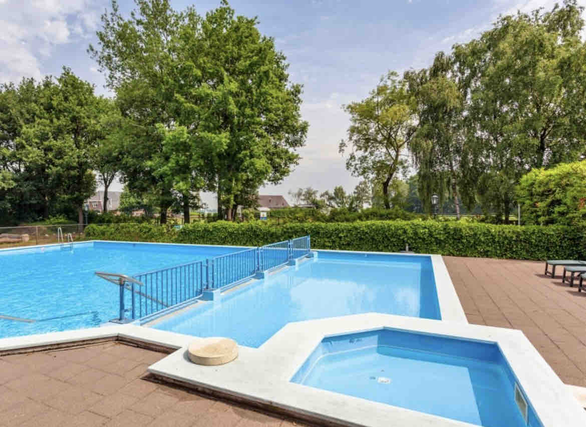 A clean outdoor swimming pool area features two pools, one larger for swimming and a smaller, circular hot tub. Surrounding greenery adds a natural touch to the serene atmosphere, complemented by a paved deck with lounge seating nearby.