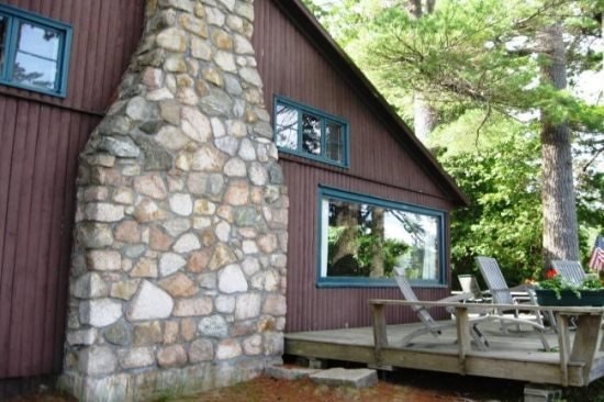 The exterior of a rustic lodge is showcased, featuring a prominent stone chimney connected to wooden siding. A spacious deck with multiple chairs is positioned in front, surrounded by lush greenery. Large windows allow natural light to fill the interior, offering a view of the outdoors.