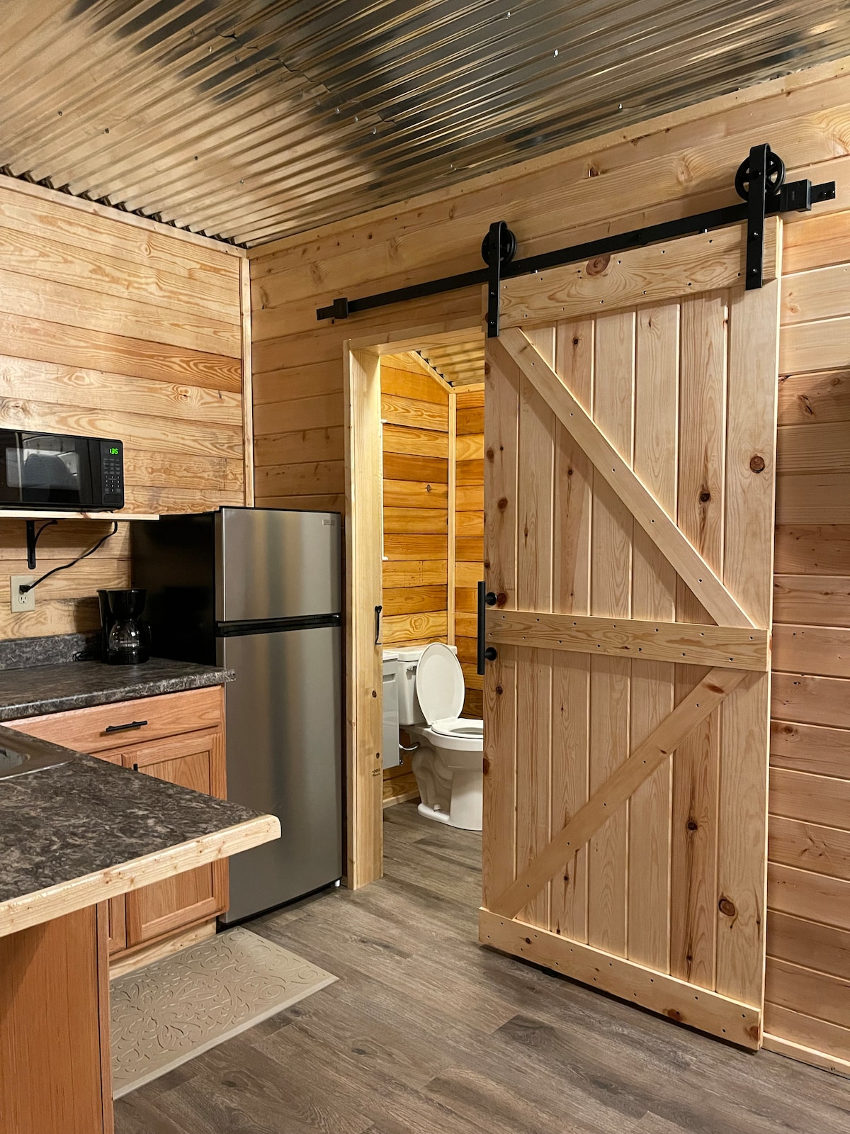 A rustic kitchen area features wooden cabinetry and a modern stainless steel refrigerator. A sliding barn door separates the kitchen from a compact bathroom, which includes a toilet. The space is illuminated by warm lighting against the pine tongue and groove walls.