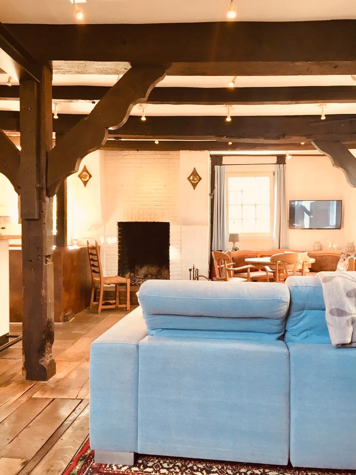 A spacious living area features a light blue sectional sofa and exposed wooden beams above. An open fireplace is located on one wall, while a dining area with wooden chairs can be seen in the background. Natural light enters through large windows adorned with light curtains.