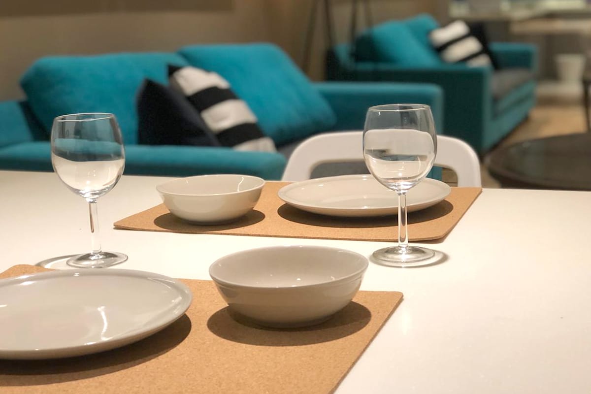 A dining area is displayed with a light-colored table featuring placemats. Two wine glasses are positioned next to a set of white plates and bowls, creating an inviting setting. The background includes a blue sofa adorned with black and white striped cushions.