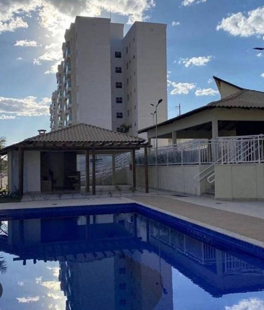 A clear view of a well-maintained swimming pool reflects the sky, surrounded by a clean patio area. A gazebo is visible on the left, with a shaded seating area. The building's modern architecture stands in the background under a partly cloudy sky.