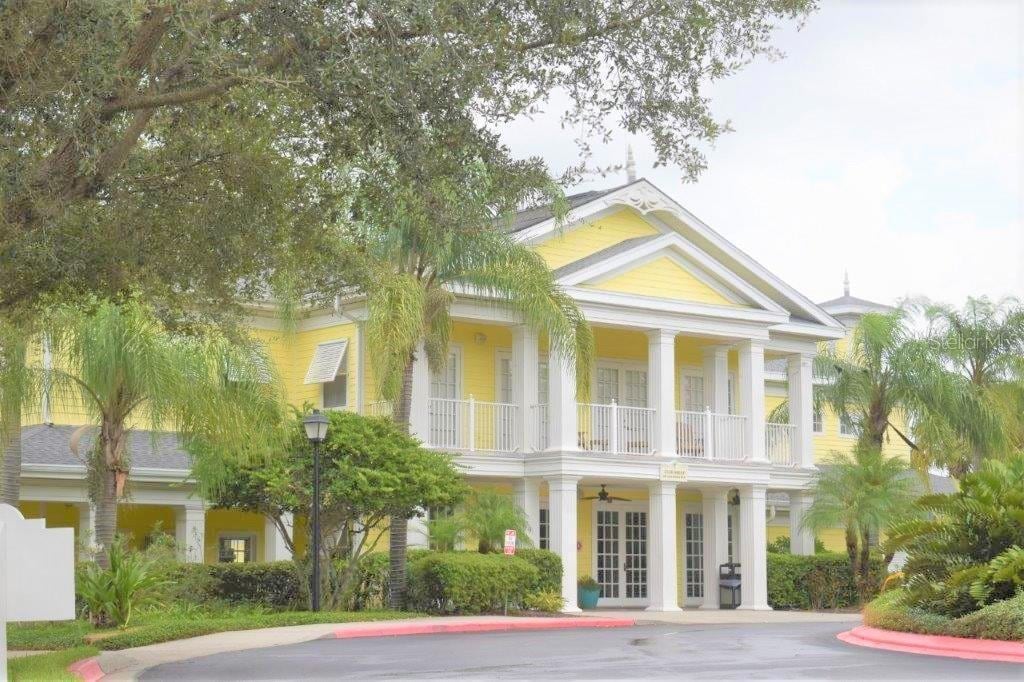 The exterior of a two-story yellow building is framed by lush greenery and palm trees. A covered porch with white railings extends across the second floor, providing an inviting entryway. The pathway to the entrance is paved, creating a welcoming approach.