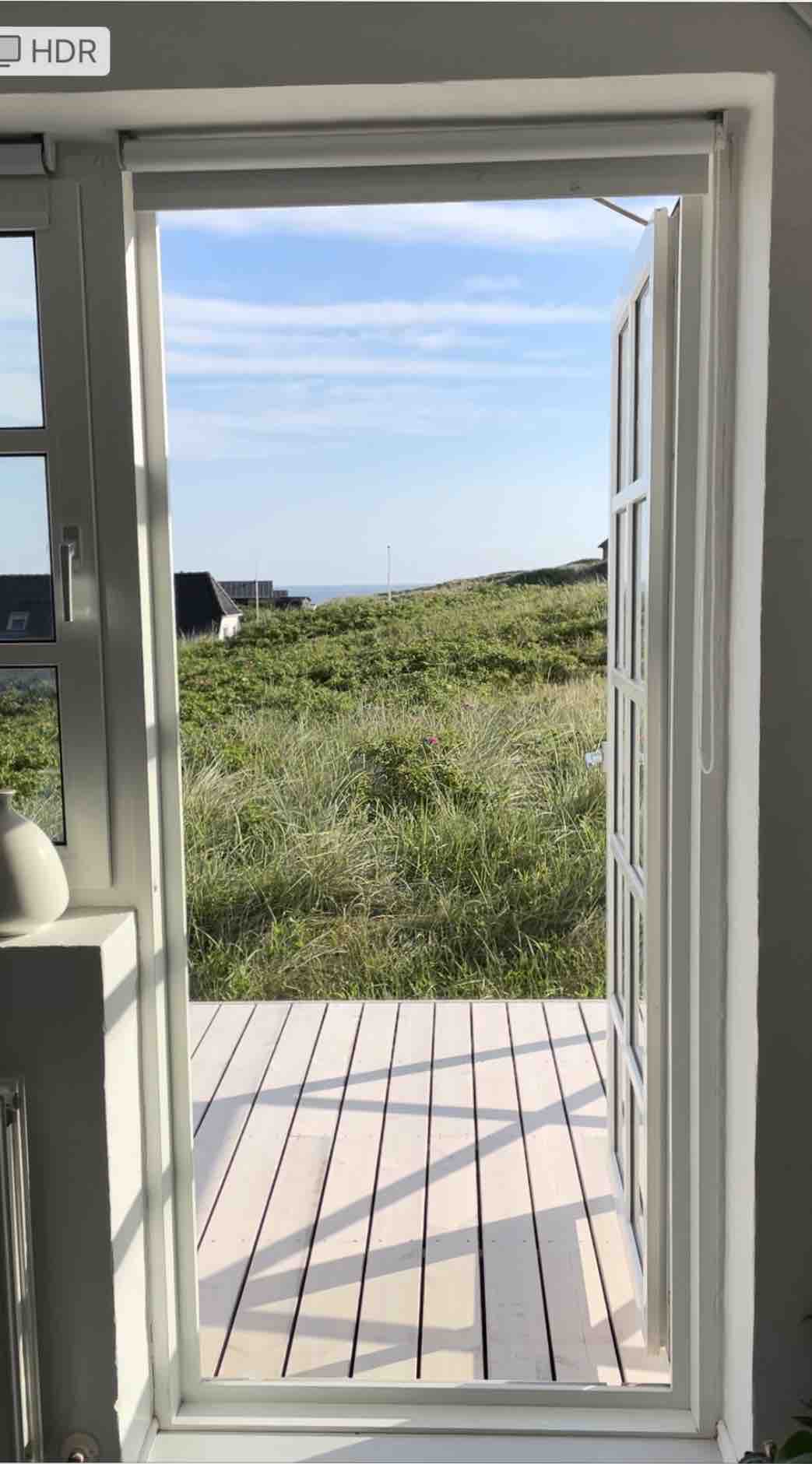 A doorway opens to a view of lush greenery and coastal landscape. The white frame contrasts with the natural surroundings, while wooden decking extends outside, inviting guests to enjoy the fresh air. Light enters the space, contributing to a bright and open ambiance.