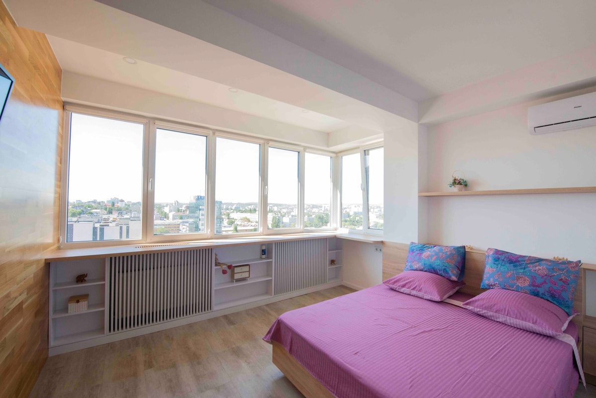 A bright sleeping area features a spacious bed dressed in lilac linens. Large windows line one wall, providing ample natural light and offering city views. Light wood accents on the walls and shelves contribute to a modern and clean design.