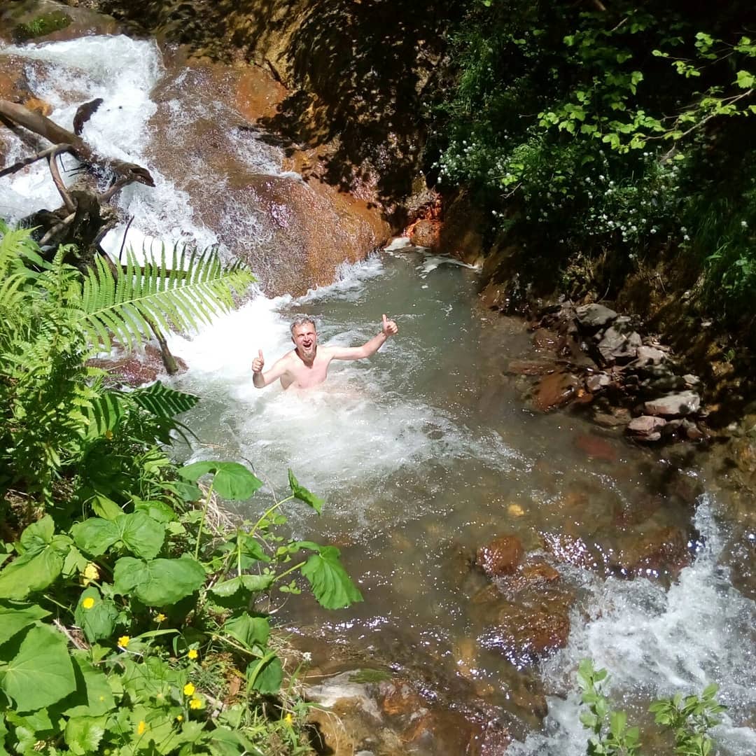 A serene natural setting is depicted, featuring clear water flowing over rocks and creating small cascades. Lush greenery surrounds the water, with ferns and vegetation framing the scene, suggesting a refreshing environment suitable for swimming and relaxation.